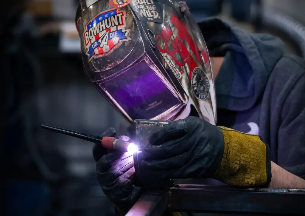 Welder using a welding torch, wearing protective gear and a custom helmet with "Bowhunt or Die" logo.